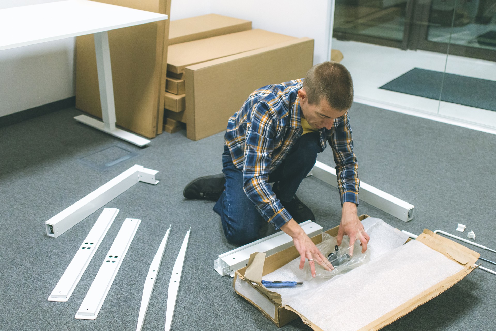Mid adult craftsman unboxing furniture parts from cardboard boxes on the floor inside modern office workspace.