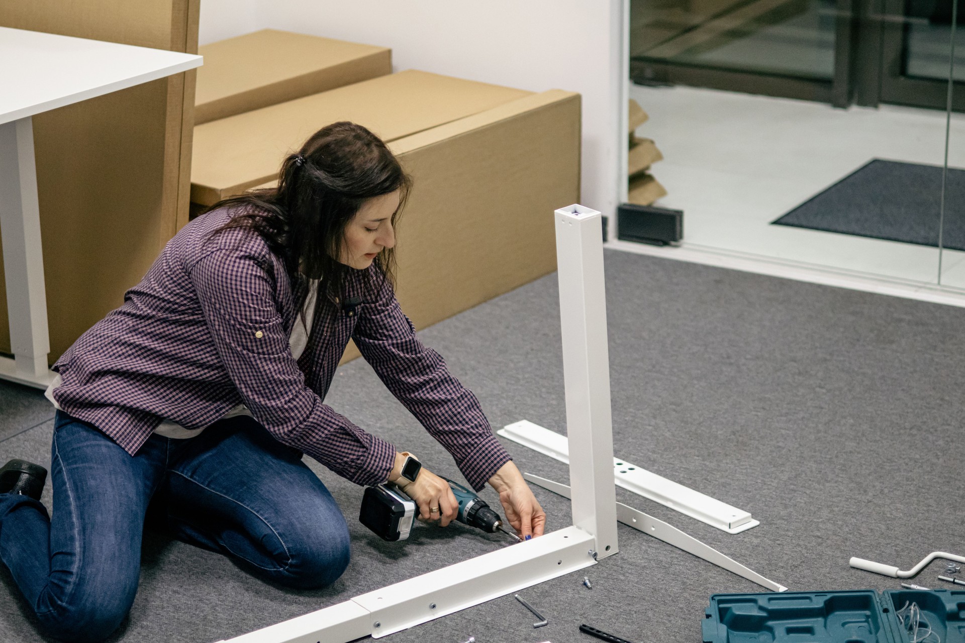Female carpenter assembling furniture on the floor inside modern office  workspace, using electric cordless power tools.